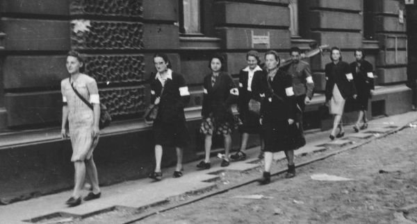 Sukienki, spódnice, bluzeczki z białym kołnierzykiem, do tego opaska na ramię. Na zdjęciu wymarsz patrolu sanitarnego Wojskowej Służby Kobiet AK na ulicy Moniuszki 9, dnia 5 sierpnia 1944 . 